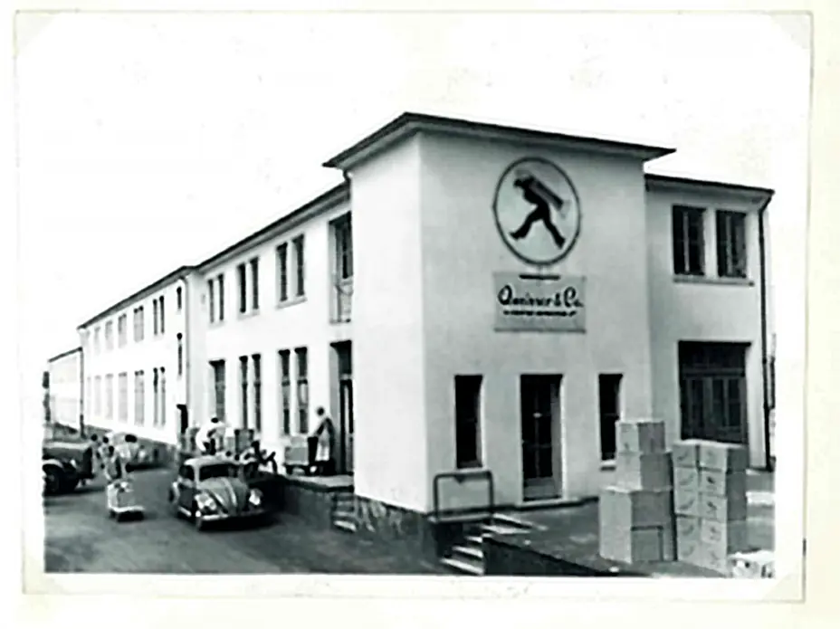 A black-and-white photograph of a building with a sign featuring a figure walking. The structure has multiple windows and a staircase leading to the entrance. Several people are gathered outside, and there are boxes stacked nearby. Vintage cars are parked in front.