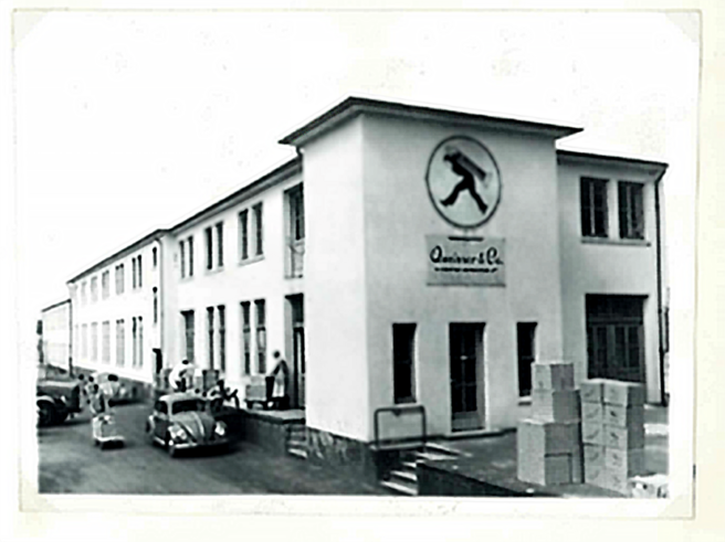 A black-and-white photograph of a building with a sign featuring a figure walking. The structure has multiple windows and a staircase leading to the entrance. Several people are gathered outside, and there are boxes stacked nearby. Vintage cars are parked in front.