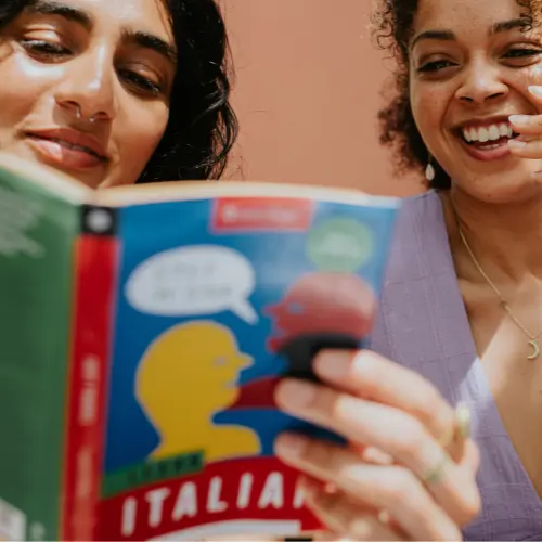 Two women engage happily while reading a colorful book titled "Learn Italian." One woman holds the book open, displaying illustrations, while the other smiles and looks on attentively. The background is a soft, solid color, contributing to a warm and inviting atmosphere.