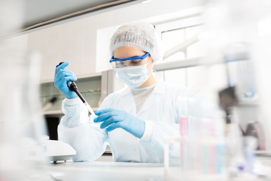A person in a lab coat, gloves, face mask, and goggles is using a micropipette in a laboratory setting. They are focused on their task, surrounded by scientific equipment and glassware.