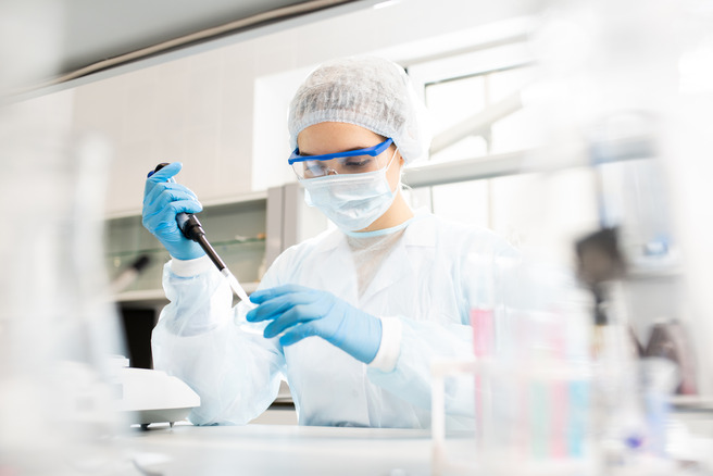 A person in a lab coat, gloves, face mask, and goggles is using a micropipette in a laboratory setting. They are focused on their task, surrounded by scientific equipment and glassware.