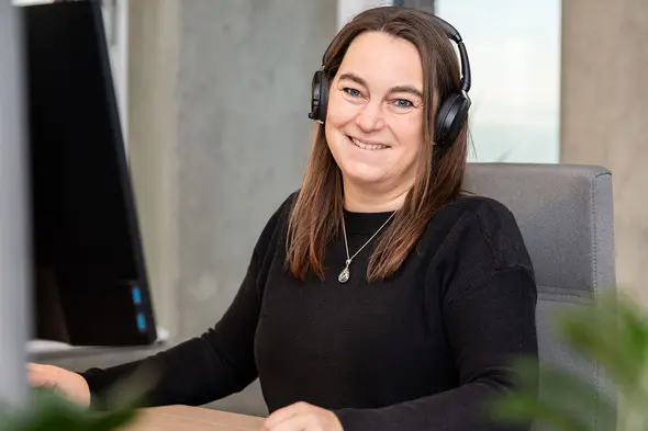 A woman with long brown hair wearing a black sweater and headphones sits at a desk, smiling at the camera. A computer monitor is visible in the background, along with some greenery.