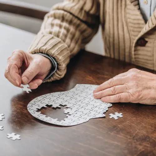 An elderly person is assembling a jigsaw puzzle on a wooden table. They are placing a puzzle piece into an almost completed section of the puzzle, which features a blank design. The focus is on their hands and the surrounding puzzle pieces scattered on the table.