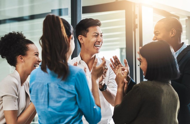 A diverse group of six people is engaged in a lively conversation, smiling and laughing together. They are dressed in casual business attire and are gathered in a bright, modern office space. The atmosphere appears joyful and collaborative.