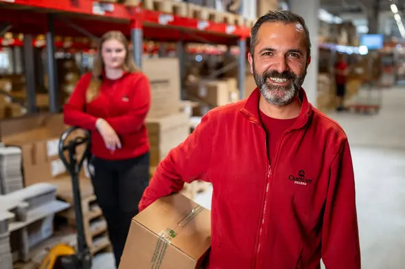 A smiling man in a red jacket holds a cardboard box in a warehouse, while a woman in the background stands near a pallet jack. The environment is filled with stacked boxes and shelves, indicating a busy shipping or storage area.