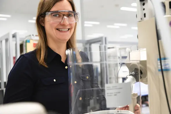 A woman wearing safety glasses and a dark shirt smiles while standing next to a laboratory machine. The background shows a professional lab environment with shelves and equipment.