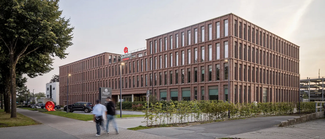 A modern brick building with multiple windows, surrounded by greenery and a sidewalk. Two people walk on the path in front, while cars are parked nearby. A large red sculpture is visible in the foreground, adding a pop of color to the urban landscape.
