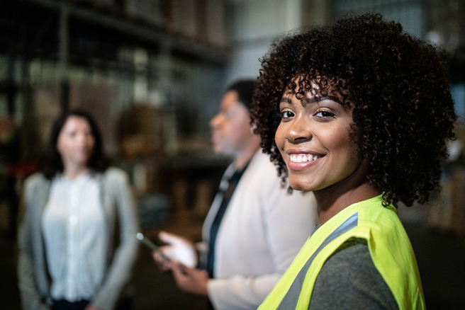 A smiling woman with curly hair is in the foreground, wearing a high-visibility yellow vest. She is standing near two other women in a warehouse setting. The background shows shelves and boxes, emphasizing an industrial environment.