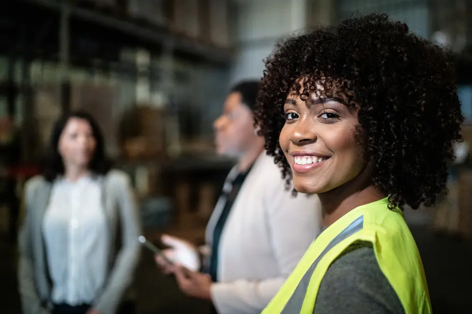 A smiling woman with curly hair is in the foreground, wearing a high-visibility yellow vest. She is standing near two other women in a warehouse setting. The background shows shelves and boxes, emphasizing an industrial environment.
