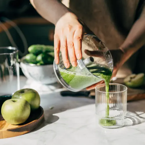 A hand pours green smoothie from a blender into a clear glass on a countertop. In the background, fresh cucumbers and green apples are visible, emphasizing a healthy, vibrant atmosphere.