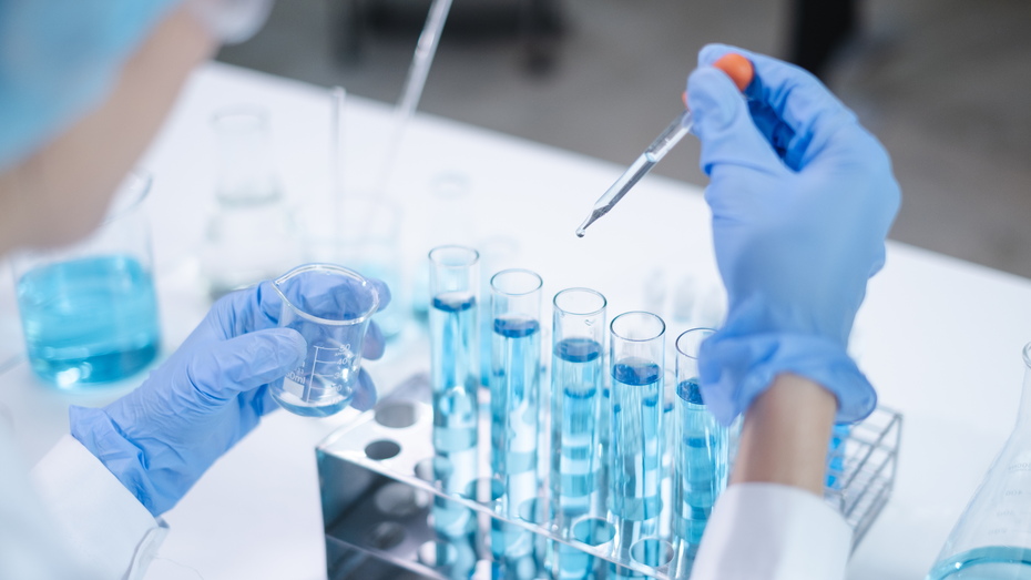 A scientist in a lab coat and blue gloves is holding a small beaker in one hand and a dropper in the other, preparing to add a liquid to a rack filled with test tubes containing blue liquid. Various lab equipment is visible in the background.