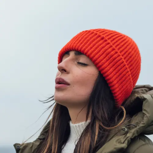 A woman with long dark hair is wearing an orange knitted beanie and a green jacket. She appears to be outdoors, with her eyes closed and a serene expression, suggesting a moment of peace or contemplation. The background is blurred and neutral, emphasizing her presence.