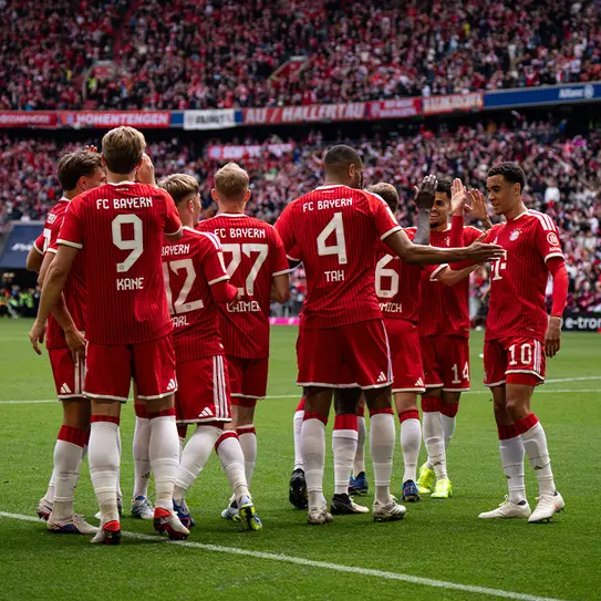 A group of FC Bayern Munich players in red jerseys huddles together on the field, celebrating and strategizing during a match, with a large crowd in the background. The atmosphere appears energetic and focused.