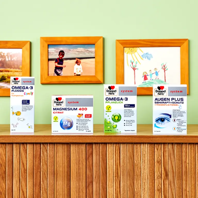 A wooden shelf displays four health supplement boxes: Omega-3 Liquid, Magnesium 400 Citrate, Omega-3 Plant-Based, and Augen Plus Eye Protection. In the background, vibrant children's drawings and photos add a colorful touch to a green wall.