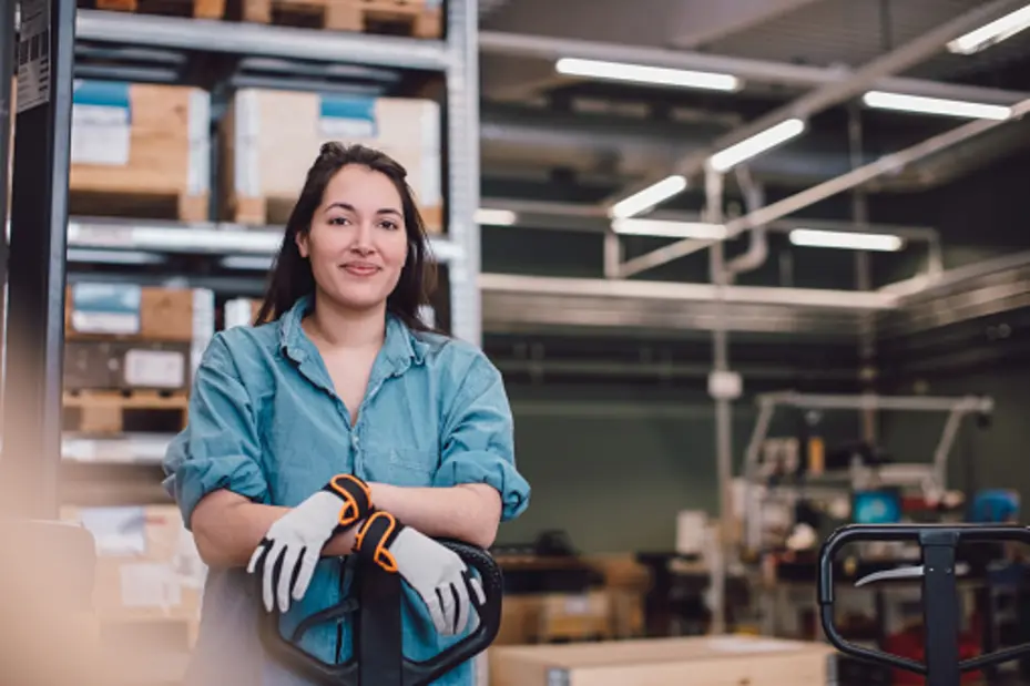 A person wearing a denim shirt and work gloves stands confidently with their arms crossed, smiling at the camera. The background features shelves with wooden crates and a workspace setting, suggesting a warehouse or industrial environment.