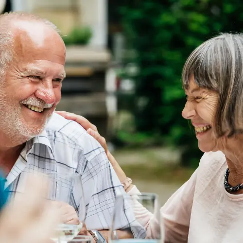 A joyful elderly couple shares a tender moment at a gathering, smiling and laughing together. The man has a gray beard and wears a striped shirt, while the woman has short gray hair and a light-colored top. Their expressions convey warmth and connection.