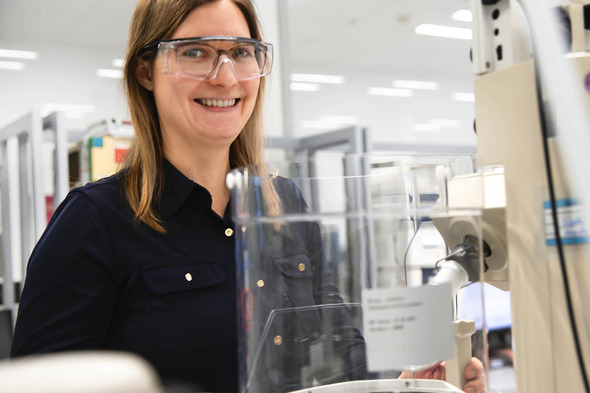 A woman wearing safety glasses and a dark shirt smiles while standing next to a laboratory machine. The background shows a professional lab environment with shelves and equipment.
