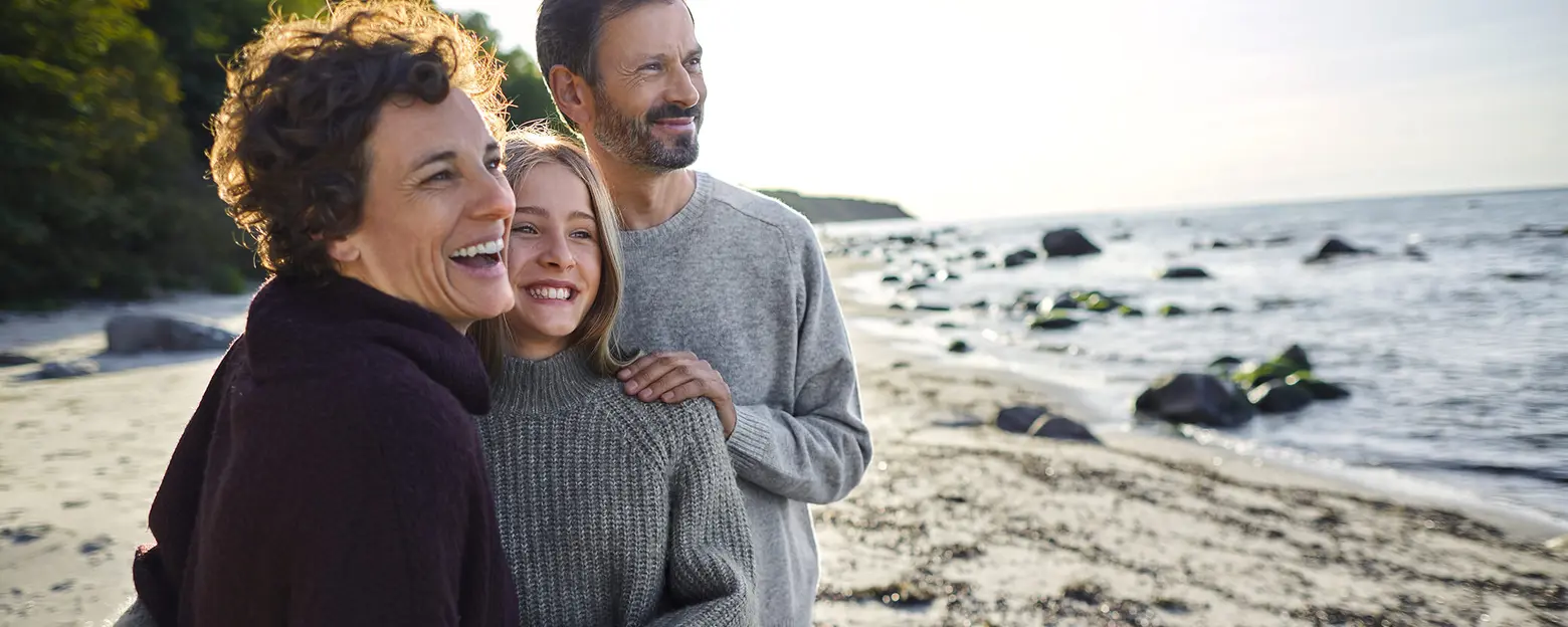 Three people stand together on a beach, smiling and enjoying each other's company. The scene features a gentle shoreline with rocks and greenery in the background, under a bright sky. The atmosphere is joyful and relaxed, capturing a moment of family togetherness.